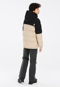 Giacca imbottita beige e nera con cappuccio, con trapuntatura orizzontale e polsini elastici. Abbinata a pantaloni grigi e scarpe nere robuste.