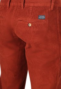 Roodkleurige corduroy broek met een knoopzak aan de achterkant en een klein gemerkt stoffen label boven de zak.