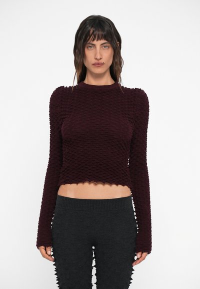 Top cropped texturat burgundy cu mâneci lungi, având un tiv cu margini festive, asortat cu pantaloni negri, riglați, cu talie înaltă, de asemenea texturați.