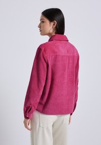 Camicia di velluto a coste fucsia con colletto, maniche con polsino a bottoni e texture a coste, mostrata di spalle con pantaloni chiari.