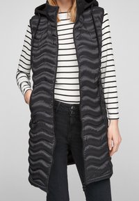 Schwarze gepolsterte Weste mit Kapuze, die eine wellenförmige Textur aufweist, mit Reißverschluss und seitlichen Taschen, über einem gestreiften Langarmoberteil getragen.