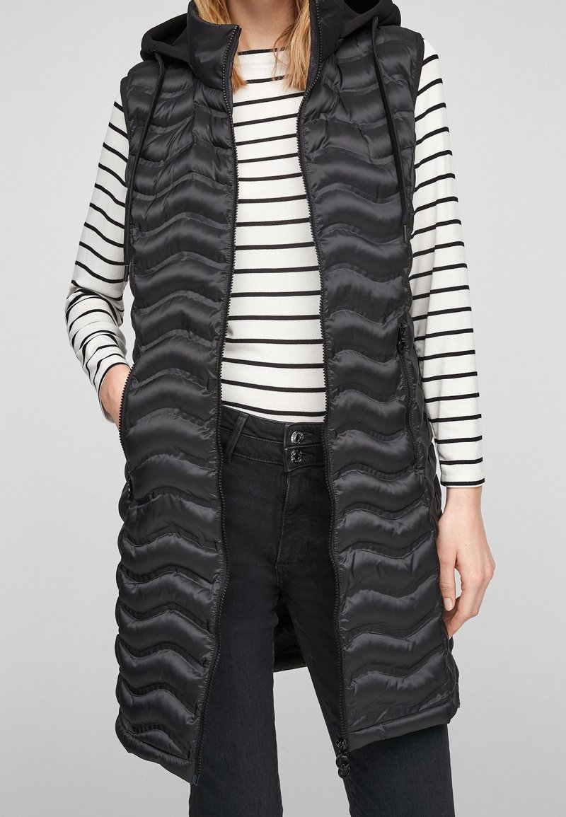 Schwarze gepolsterte Weste mit Kapuze, die eine wellenförmige Textur aufweist, mit Reißverschluss und seitlichen Taschen, über einem gestreiften Langarmoberteil getragen.