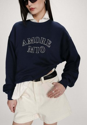 Femme portant un sweat-shirt bleu marine avec le texte "AMORE MIO", une chemise blanche à col en dessous, un short crème, une ceinture noire, des bagues et des lunettes de soleil noires.