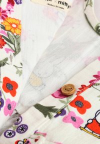 Tissu léger à motif floral avec des fleurs multicolores ; présente un bouton en bois et des coutures visibles. Fond blanc.