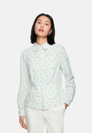 Frau mit glatten dunklen Haaren, die ein weißes Hemd mit grünem Rautenmuster und weiße Hosen trägt, posiert mit einer Hand in der Tasche.