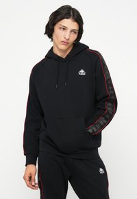 Sudadera con capucha negra y costuras en rojo. Cuenta con un bolsillo tipo canguro y un logo en el pecho. Los paneles laterales tienen un diseño estampado.