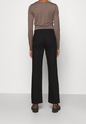 Personne de dos portant une chemise taupe à manches longues, un pantalon large noir et des chaussures à plateforme marron sur un fond uni.