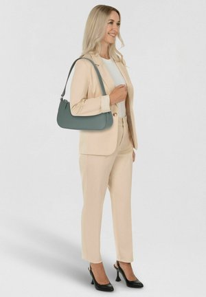 Mujer con traje beige y top blanco, llevando un bolso verde azulado sobre el hombro, usando zapatos de tacón alto negros con tira trasera, sonriendo.