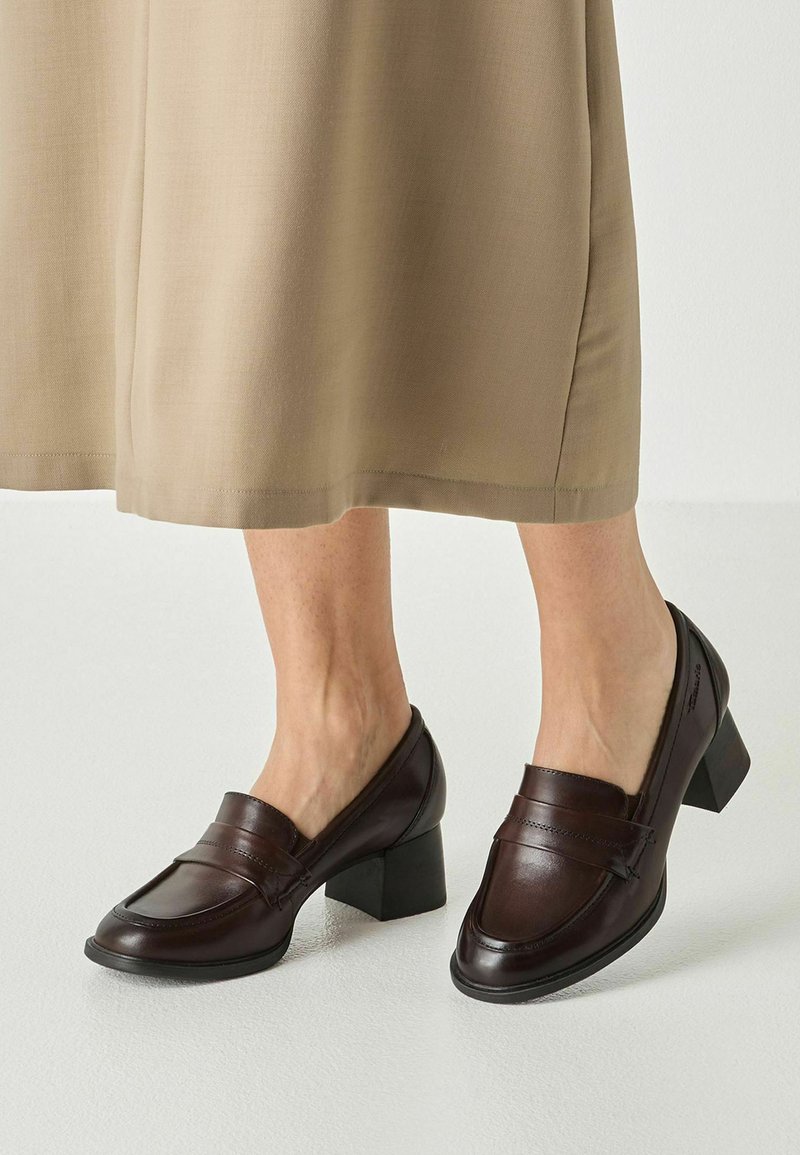 Jambes portant une jupe beige et des mocassins en cuir marron foncé à talons blocs, debout sur un sol clair devant un fond blanc.