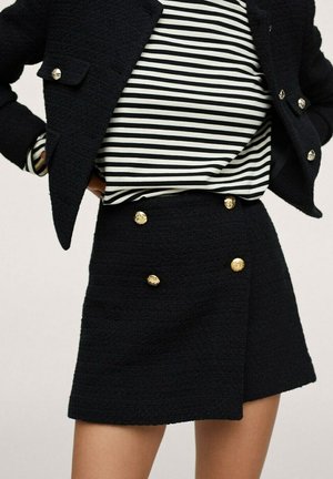 Vrouw draagt zwarte textuurrijke rok met gouden knopen, zwart textuurrijk kort jasje en zwart-wit gestreept topje met lange mouwen.
