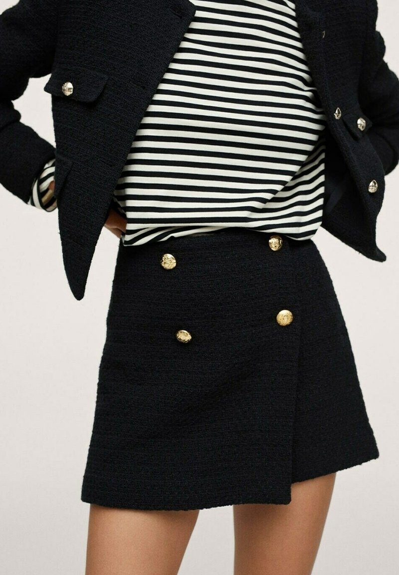 Femme portant une jupe texturée noire avec des boutons dorés, une veste courte texturée noire, et un haut à manches longues rayé noir et blanc.