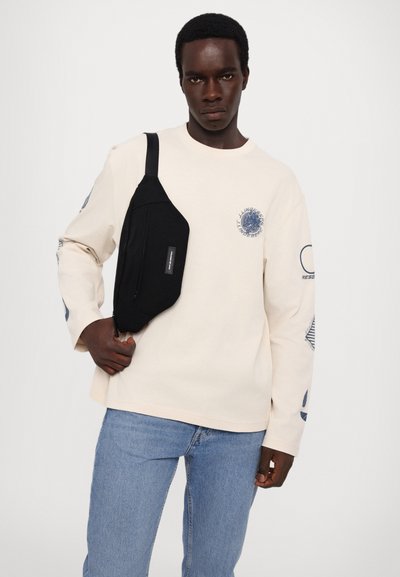 Light beige long-sleeve t-shirt with circular graphic on chest and blue accents, paired with blue jeans and a black crossbody bag.
