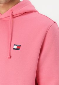Roza dukserica iz mehkega materiala, z sprednjim žepom in kapuco na vrvico. Prikazuje majhen logotip z blagovno znamko "TOMMY JEANS".