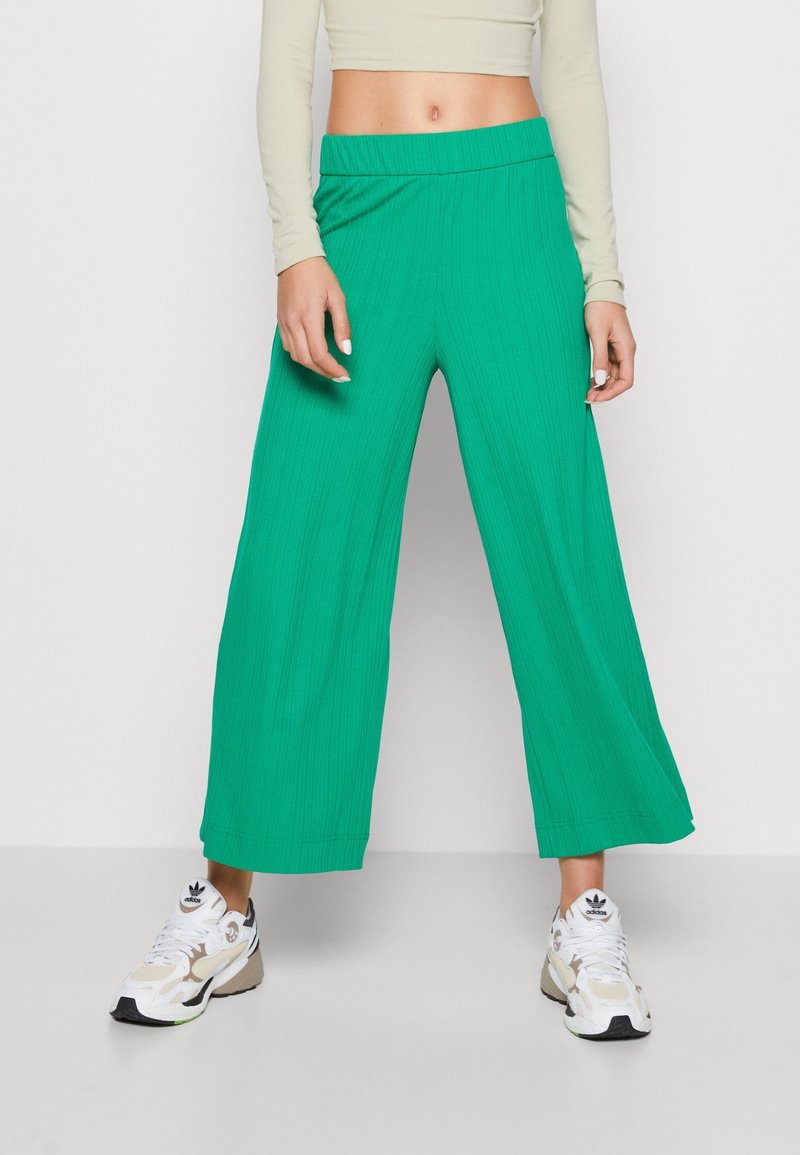 Femme portant un pantalon large vert vif, un haut beige à manches longues raccourci, et des baskets blanches Adidas, debout devant un fond uni.