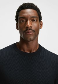 Dark blue sweater with a textured herringbone pattern, round neckline, and ribbed cuffs. Close-up of the garment on a neutral background.