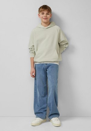 Jeune personne souriante portant un sweat à capuche beige, un jean bleu ample et des baskets blanches, debout devant un mur gris clair uni.