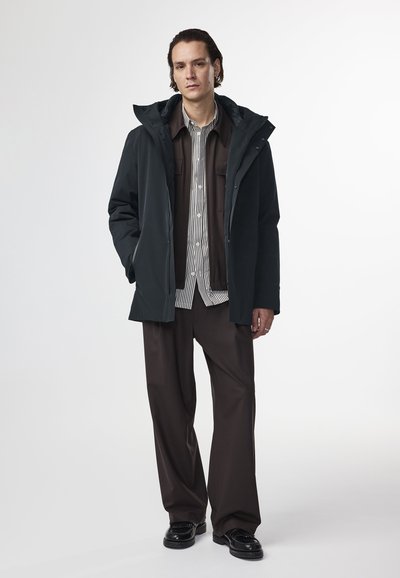 Parka verde scuro con cappuccio, chiusura con zip e tasche laterali, indossato sopra una camicia a righe e pantaloni larghi marroni. Le scarpe nere completano il look.