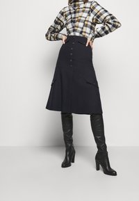 Navy A-line skirt with a high waist, featuring front button closure, two side pockets, and smooth texture, paired with a plaid shirt and tall black boots.