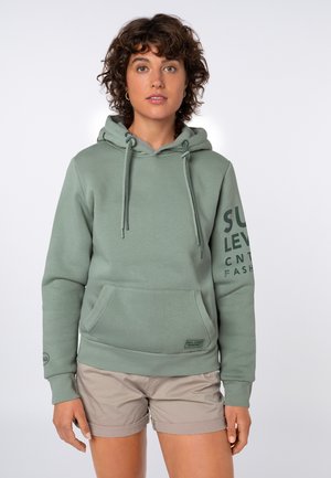 Jong persoon met krullend haar draagt een groene hoodie met voorzak en beige korte broek, staand tegen een effen lichte achtergrond.