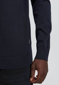 Maglione a maglia blu navy con una texture a coste, dotato di una toppa con logo laterale e polsini aderenti, mostrato su uno sfondo scuro.