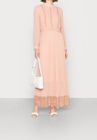 Robe longue plissée en pêche doux avec une bordure en dentelle à l'ourlet. Présente des manches longues et un col montant. Accessoirisée avec un sac à main blanc et des chaussures.