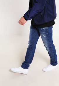 Marinjacka över en svart skjorta, slitna blå jeans och vita sneakers, med synliga sömmar och en avslappnad passform.