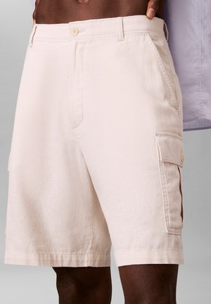 Nærbillede af en person iført lyse beige cargoshorts med knaplukning på sidelommerne og slidser foran på lommerne.