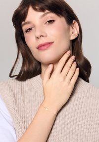 Złota bransoletka z zawieszką w kształcie litery "M", noszona na dłoni opierającej się na swetrze o beżowej fakturze, w delikatnych kolorach i naturalnym świetle.