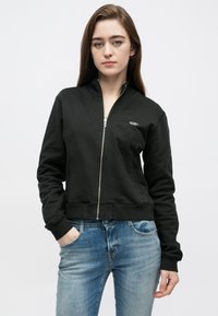 Sweatshirt preto com fecho ziper feito de algodão, com gola alta e punhos canelados, combinado com jeans azuis. Design simples e casual.
