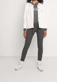 Veste en denim blanche sur un hoodie gris avec le texte "New York", associée à un pantalon noir à côtes et des baskets blanches, se tenant sur un fond neutre.