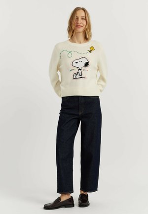 Vrouw draagt een witte trui met Snoopy- en Woodstock-ontwerp, donkerblauwe spijkerbroek en zwarte loafers, staand tegen een effen achtergrond.