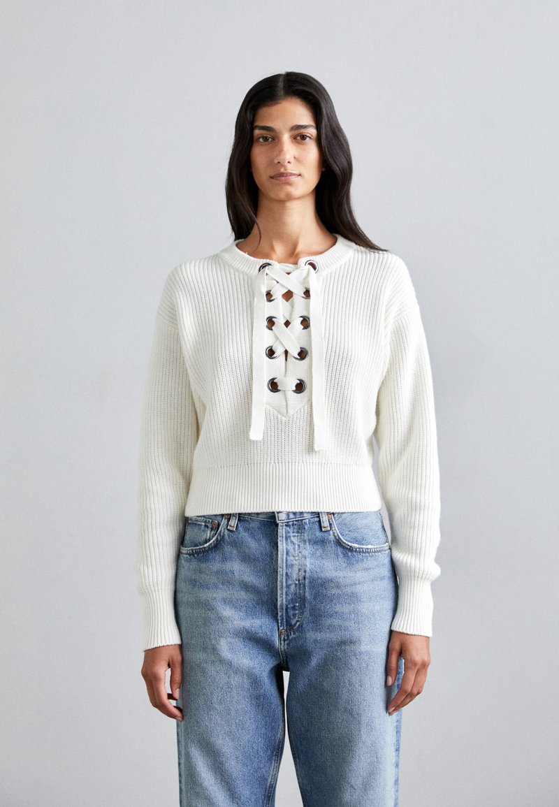 Witte ribgebreide trui met een ronde halslijn en een vetersluiting aan de voorkant, vastgezet met metalen ringetjes, gecombineerd met lichtblauwe high-waisted jeans.