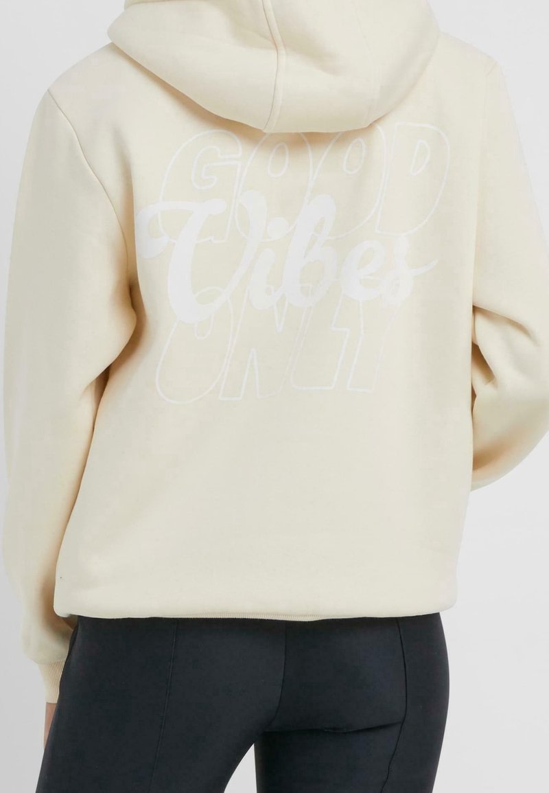 Personne portant un sweat à capuche beige avec le texte « Good Vibes Only » dans le dos, associé à un pantalon noir moulant, vue de dos.