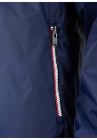 Primo piano di una giacca blu navy con una cerniera laterale con accenti rossi e bianchi, e un tessuto liscio e strutturato.