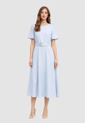 Donna in piedi che indossa un abito midi azzurro chiaro con maniche corte, scollo tondo e cintura abbinata con fibbia dorata, abbinato a tacchi beige.