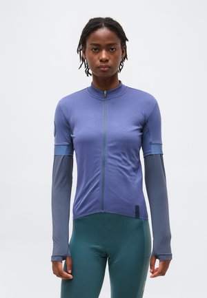 Žena s krátkymi dredmi, ktorá má oblečený priliehavý fialový cyklistický dres so zapínaním na zips cez dlhé sivé rukávy a tyrkysové cyklistické nohavice, stojaca na bielom pozadí.