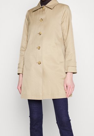 Manteau beige mi-longueur aux genoux avec cinq boutons, poches avant et détails aux poignets, porté sur un jean bleu foncé et un haut noir.