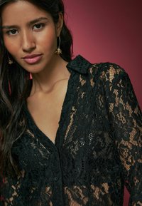 Black lace top featuring a collared neckline and button detail, showcasing floral patterns and a sheer texture against a red background.