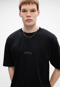 Joven vestido con una camiseta negra de HUGO y una cadena de plata, frente a un fondo blanco liso.