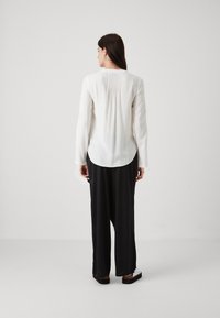 Blusa bianca a maniche lunghe con pieghe posteriori, orlo arrotondato e texture liscia. Abbinata a pantaloni neri larghi e scarpe basse.