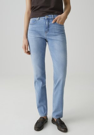 Person, die hellblaue Jeans mit geradem Bein, schwarze Loafer und ein dunkelbraunes Oberteil trägt, steht vor einem schlichten grauen Hintergrund.