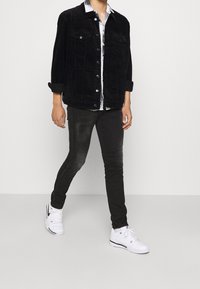 Chaqueta de pana negra con cierre de botones y dos bolsillos en el pecho, combinada con jeans ajustados negros desgastados y zapatillas blancas.