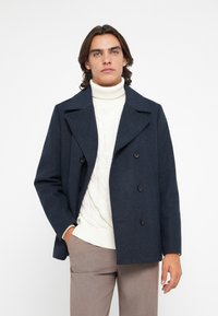 Mornarica vuna peacoat s dvostrukim kopčanjem, crnim gumbima i oštrim ovratnikom, nosi se preko kremaste džersej turtleneck majice.