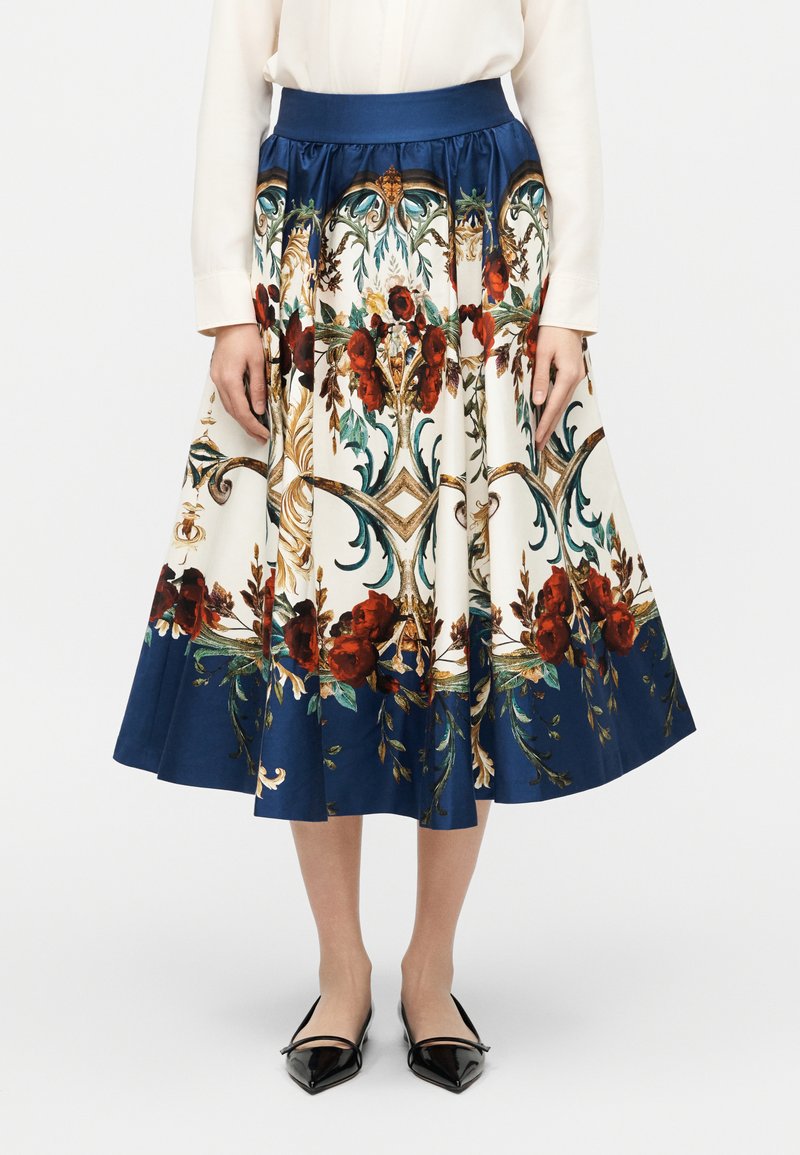 Jupe mi-longue à motif floral avec ceinture et ourlet bleu marine, portée avec un chemisier blanc et des ballerines noires à bouts pointus.