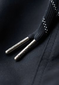 Zwarte trekkoorden met zilverkleurige metalen uiteinden bedrukt met "SuperdrySwim" op donker textiel met zichtbare stiksels.