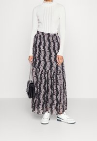Schwarz-maxi Rock mit Paisley-Muster, gestuftes Design. Kombiniert mit einem weißen gerippten Oberteil und weißen Sneakern. Hält eine schwarze Handtasche.