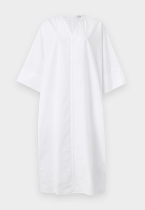 Kobieta w luźnej białej sukience midi z rękawami do łokcia, brązowych sandałach i trzymająca małą ciemnobrązową torebkę.