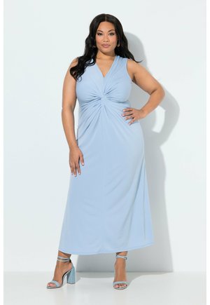 Kvinde med langt mørkt hår iført en ærmeløs lyseblå midi-kjole med tvistdetalje foran og matchende høje hæle, poserer selvsikkert.