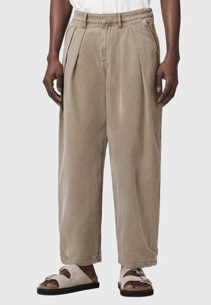 Man die losse beige broek met plooien aan de voorkant en beige sandalen met open teen en zwarte zolen draagt, staand tegen een effen achtergrond.