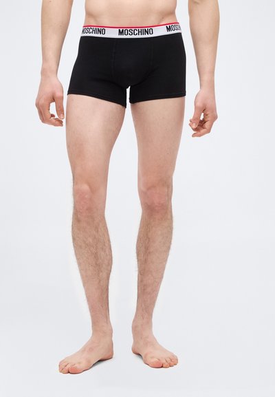 Άνδρας φοράει μαύρα μποξεράκια Moschino με λευκή ζώνη μέσης με λογότυπο, στέκεται ξυπόλητος μπροστά σε απλό ανοιχτό φόντο.